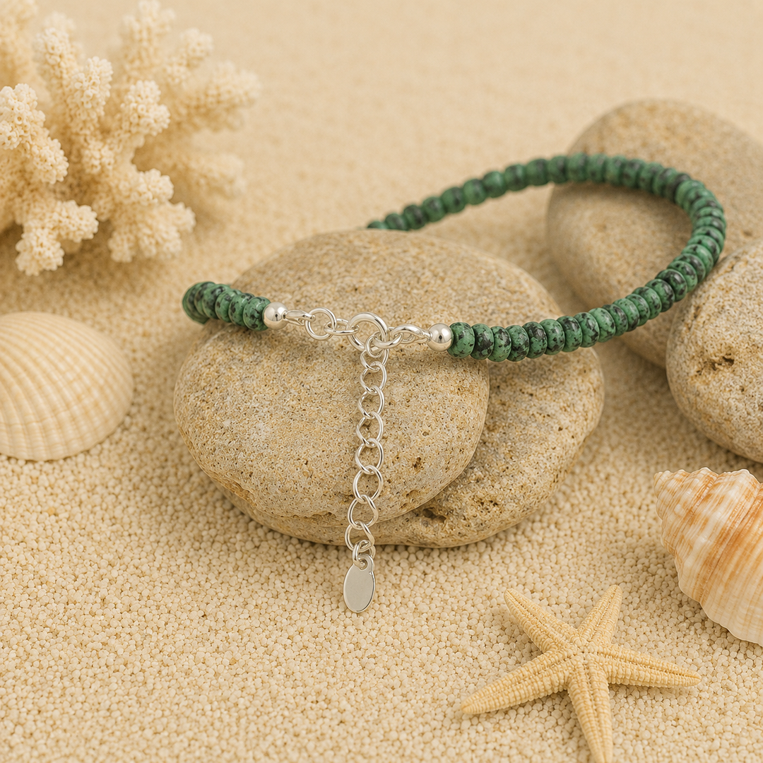 Green beaded anklet with silver chain on a sandy background with stones, shells, and starfish.