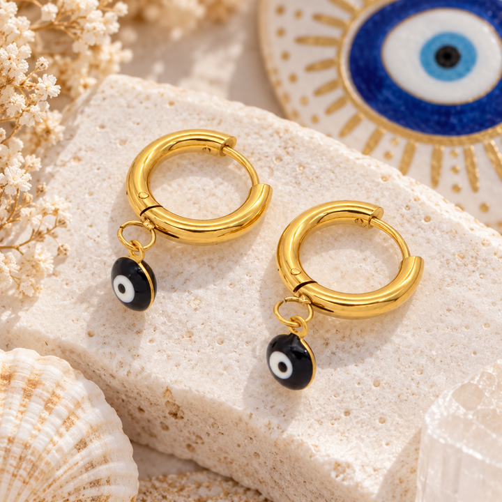 A pair of gold huggie hoop earrings with small dangling evil eye charms, styled on a textured white stone block with seashells and dried flowers.