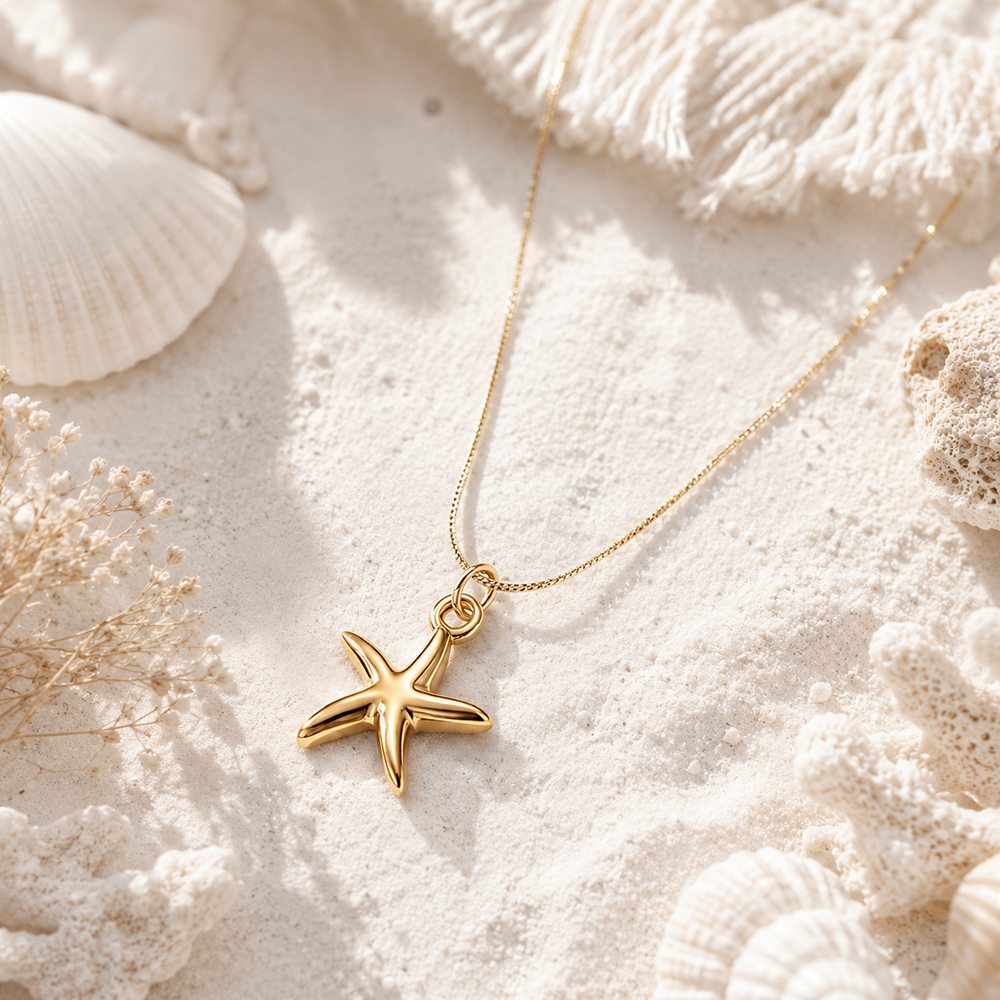 A gold starfish pendant necklace on a delicate gold chain, styled on a bed of white sand with seashells and dried flowers.