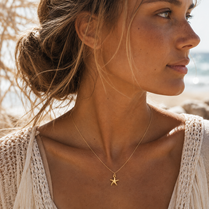 A woman in a white crochet top wearing a delicate gold necklace with a starfish pendant, photographed outdoors on a sunny beach.