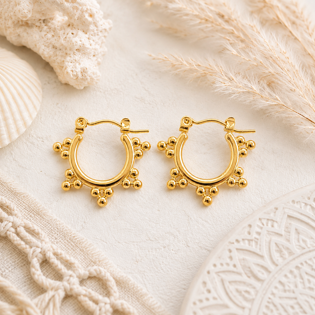 A pair of gold boho hoop earrings with small bead-like accents around the lower edge, displayed on a light cream textured surface 