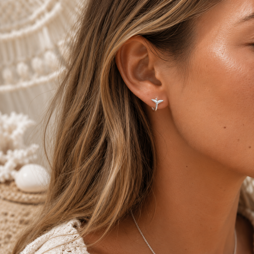 A close-up of a woman's ear wearing a small silver whale tail earring set against a warm boho-style background with a macrame wall hanging and woven textiles.