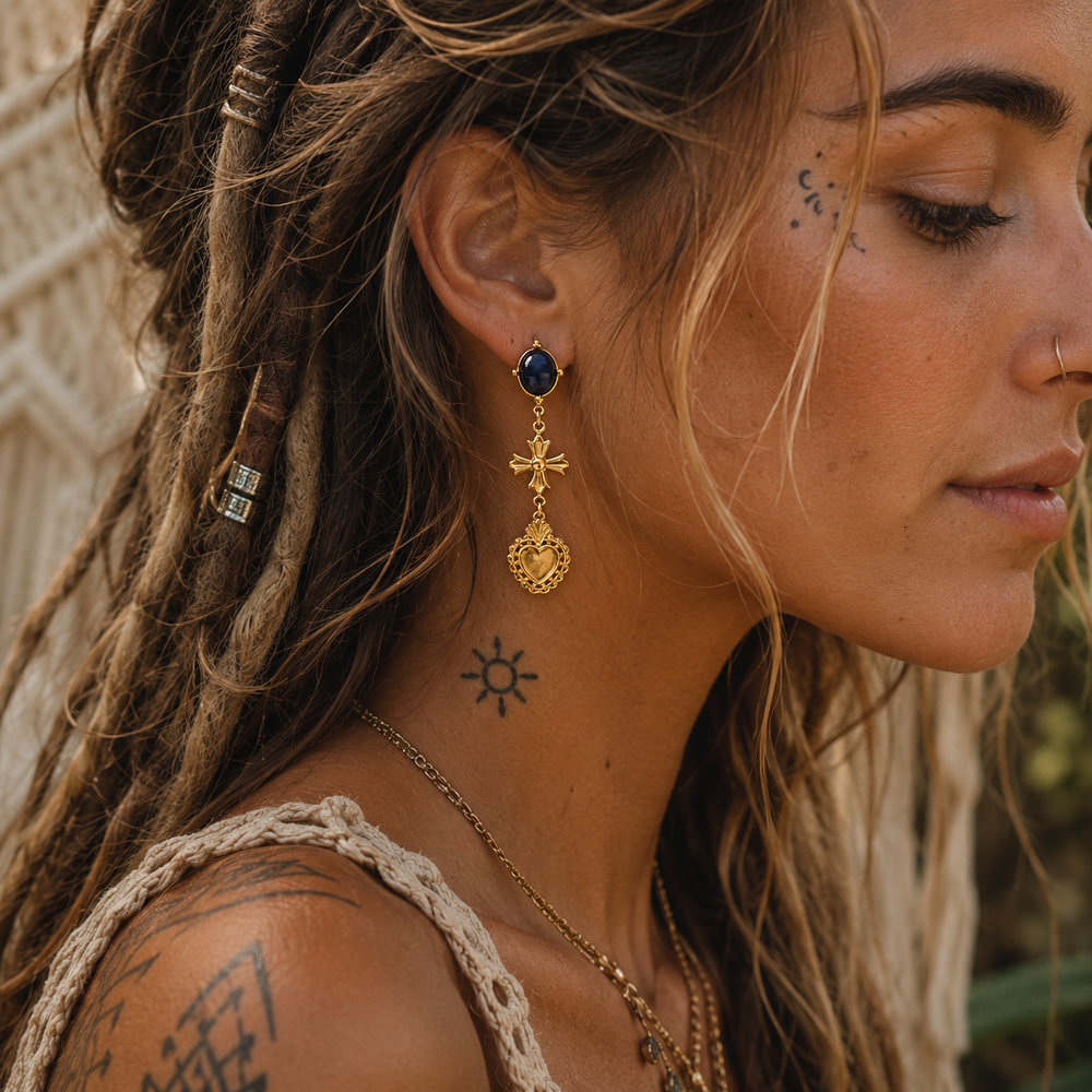 A close-up side profile of a woman with dreadlocks wearing a gold-toned bohemian-style drop earring with a dark blue oval stone and a heart-shaped charm.