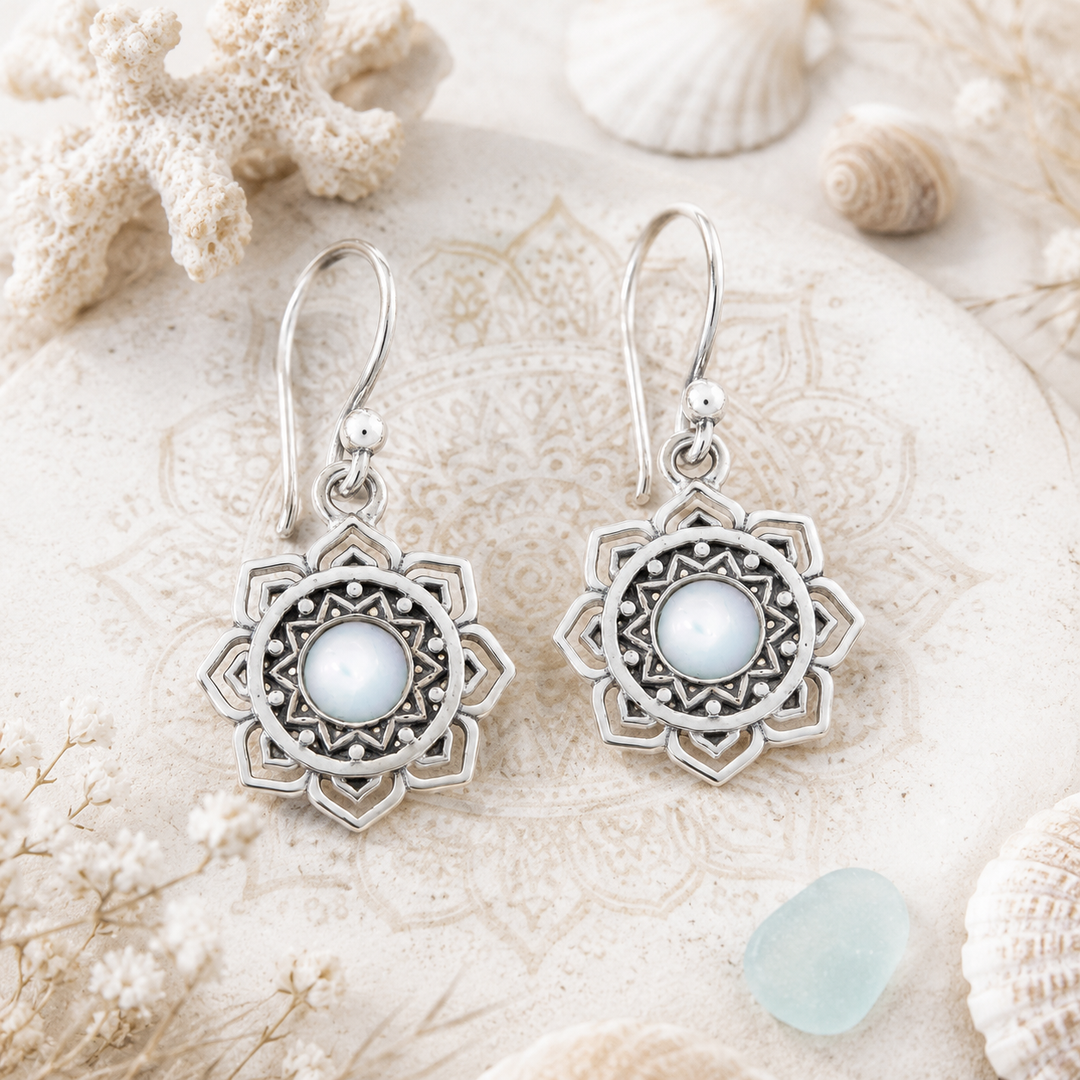 A pair of silver-toned bohemian-style dangle earrings with intricate filigree sunburst designs and central white mother-of-pearl stone displayed on a textured beige stone surface surrounded by seashells and coral.