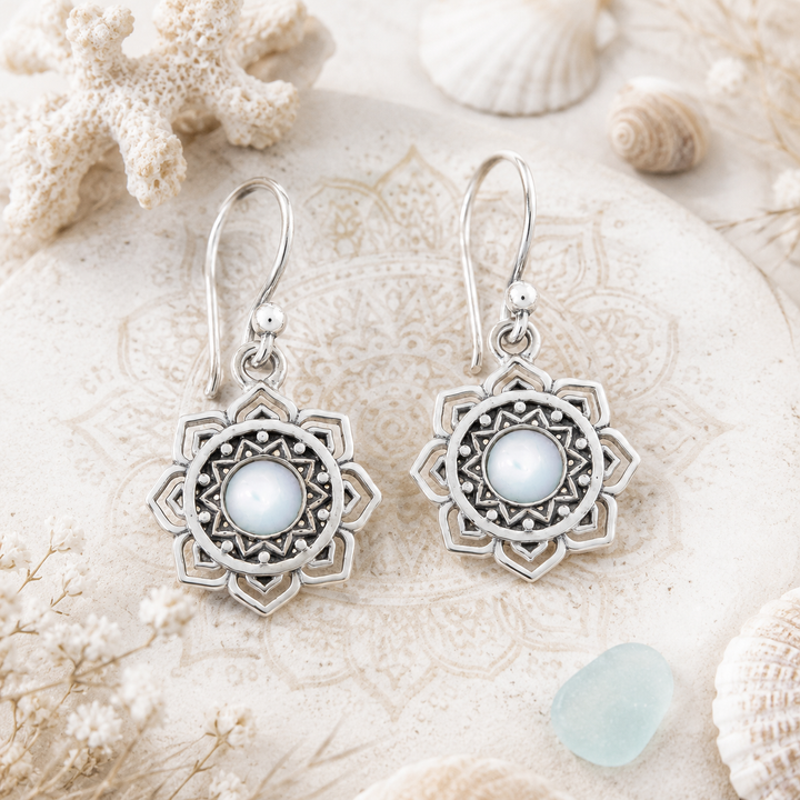 A pair of silver-toned bohemian-style dangle earrings with intricate filigree sunburst designs and central white mother-of-pearl stone displayed on a textured beige stone surface surrounded by seashells and coral.