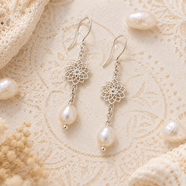 A pair of silver drop earrings with floral filigree charms and white pearls, displayed on a textured cream lace surface with scattered pearl beads.
