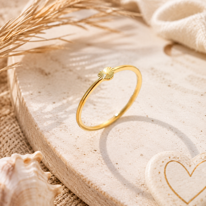 A thin gold ring with a small sunburst or fan-shaped detail on the front, displayed on a round beige stone slab with dried grass, a shell, and a heart-shaped stone nearby.