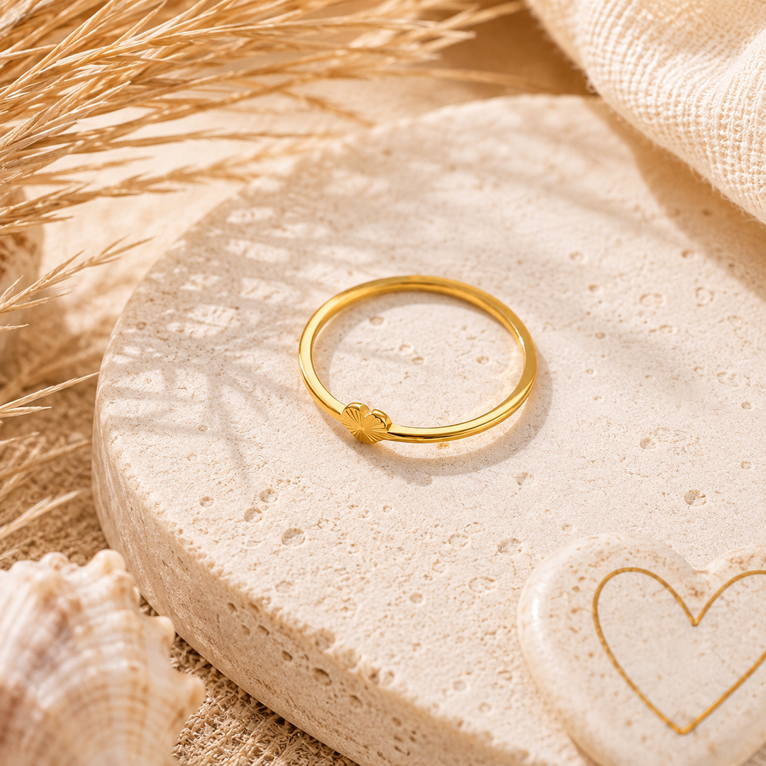 A thin gold ring with a small heart-shaped charm, displayed on a textured beige stone dish with dried grass, a seashell, and a linen cloth in the background.
