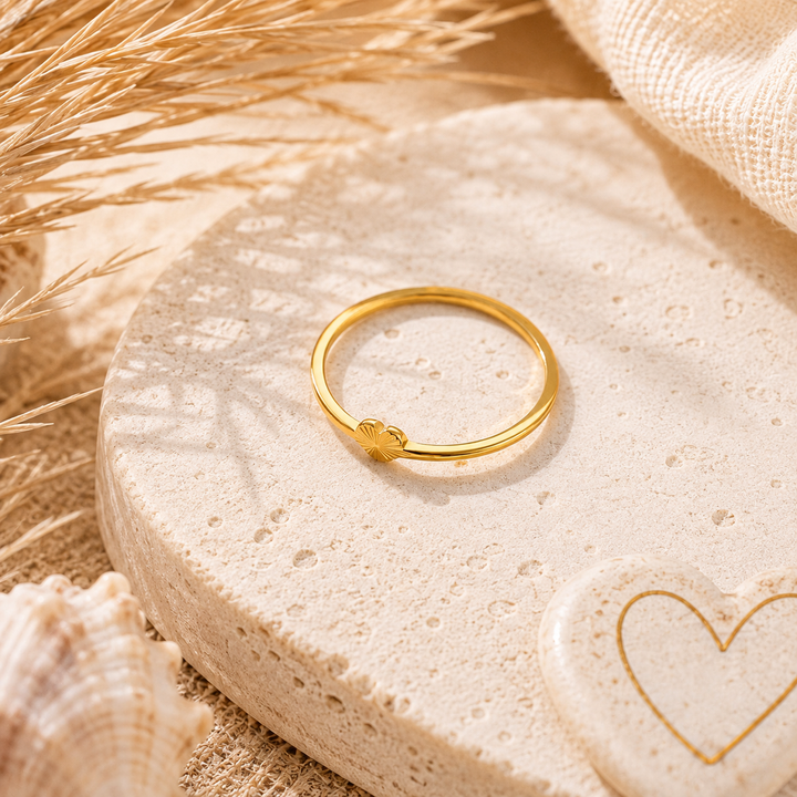 A thin gold ring with a small heart-shaped charm, displayed on a textured beige stone dish with dried grass, a seashell, and a linen cloth in the background.
