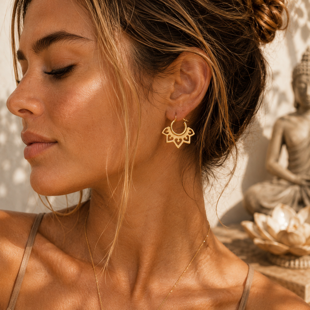 A close-up lifestyle shot of a woman wearing a delicate gold lotus-shaped hoop earring, with warm sunlight highlighting her skin and hair, set against a softly blurred background featuring a Buddha statue.