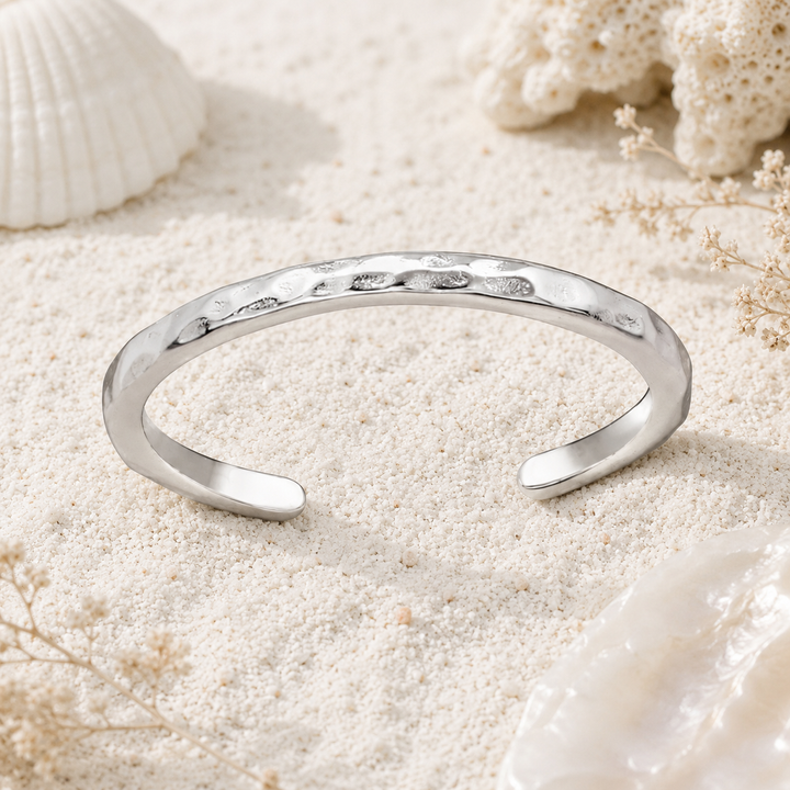 A polished silver toe ring with a hammered texture, displayed on a bed of white sand with seashells and dried flowers.