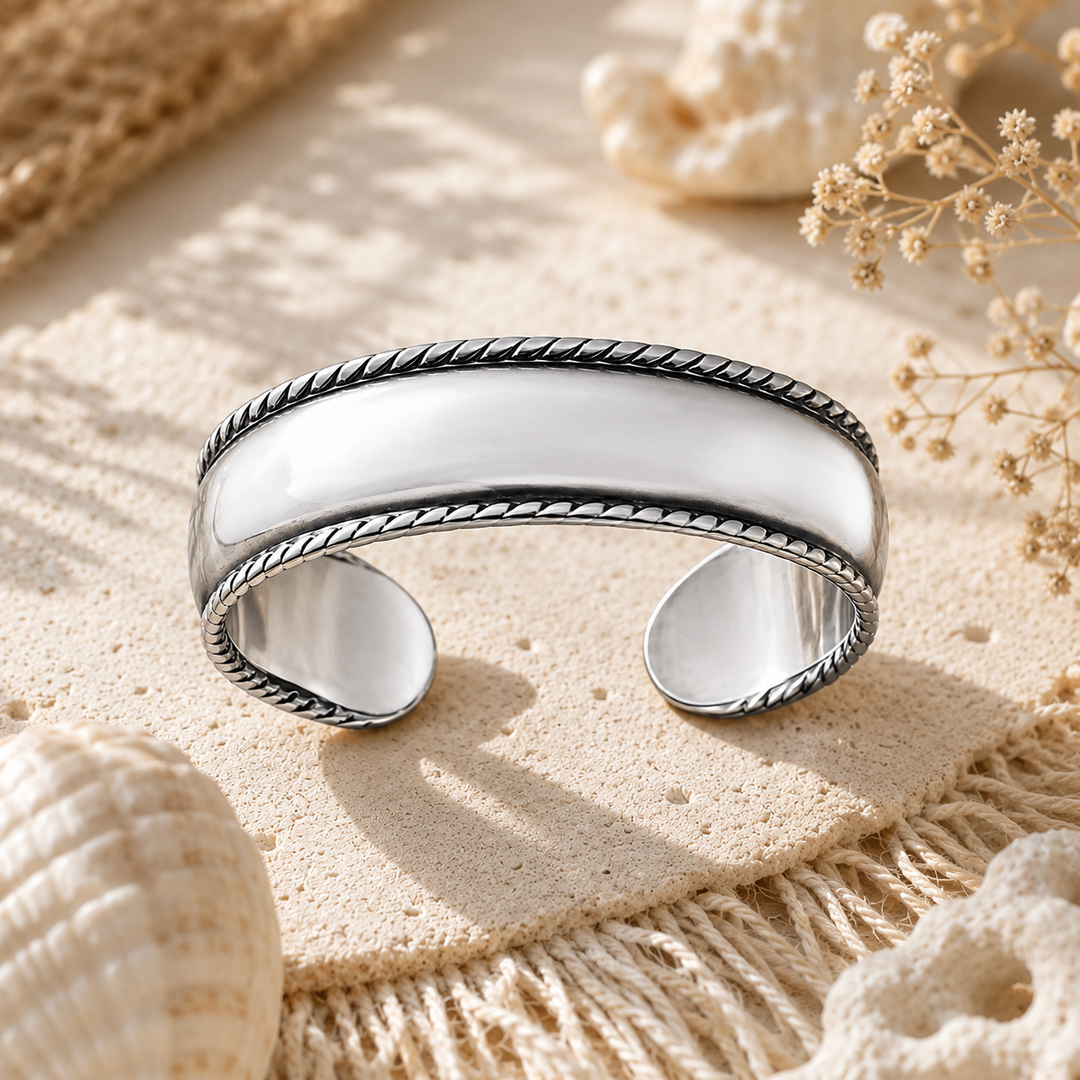 A polished sterling silver toe ring with a twisted rope border, displayed on a textured beige fabric with seashells and dried flowers.