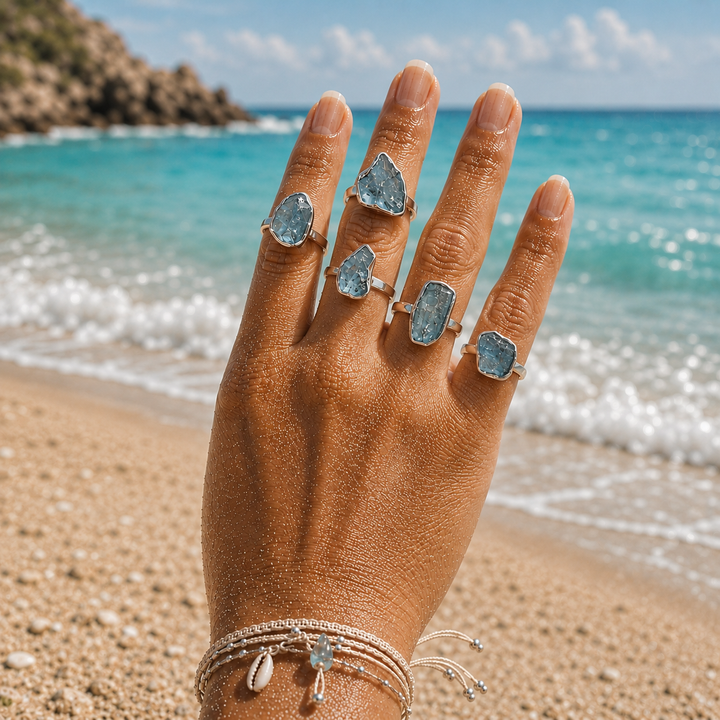 A close-up of a hand with five silver rings featuring rough, irregular-cut aquamarine rings, set against a blurred beach background with turquoise ocean water and sand.