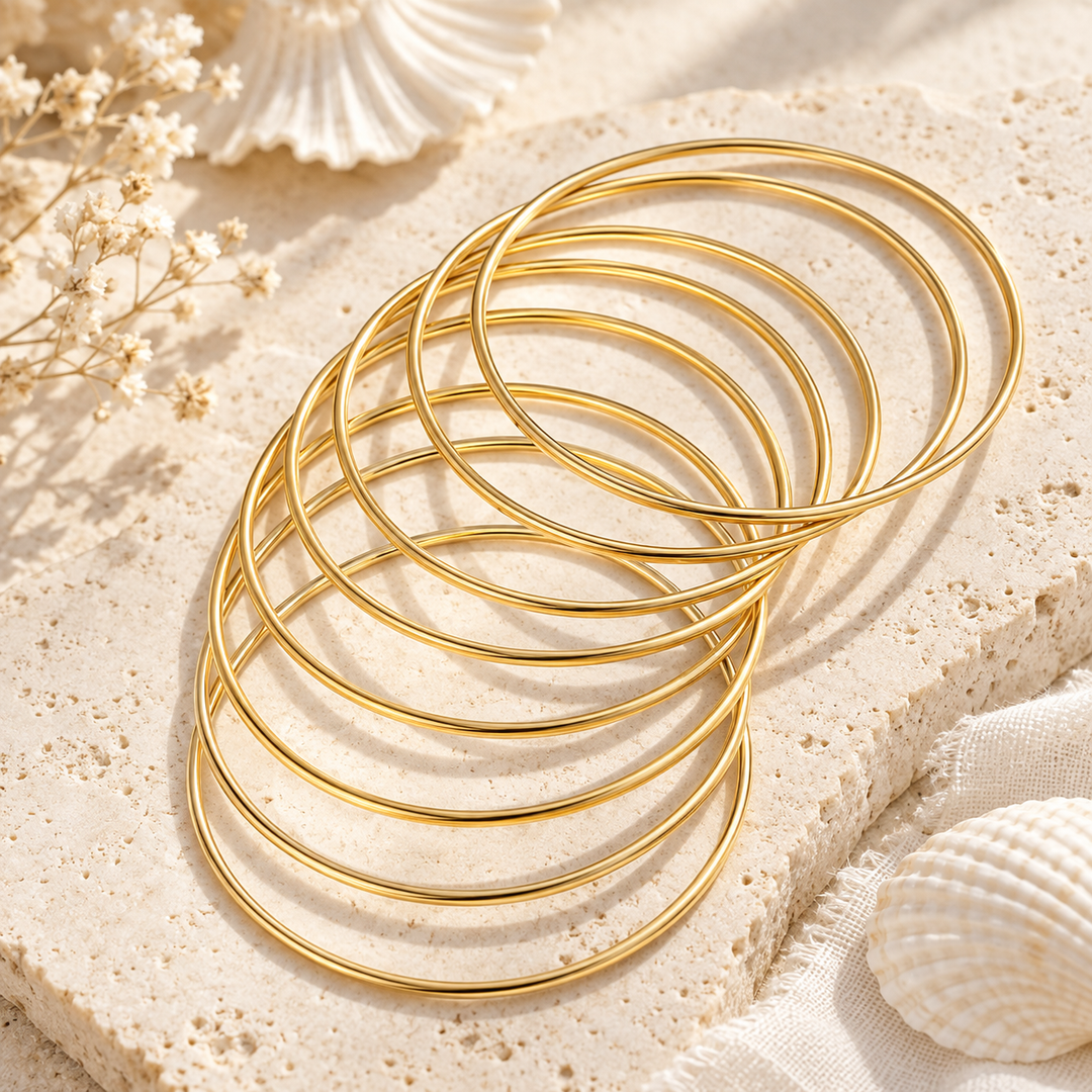 A stack of thin, polished gold bangles or rings arranged in an overlapping fan shape on a textured beige stone slab, with small dried flowers and seashell props in the background.