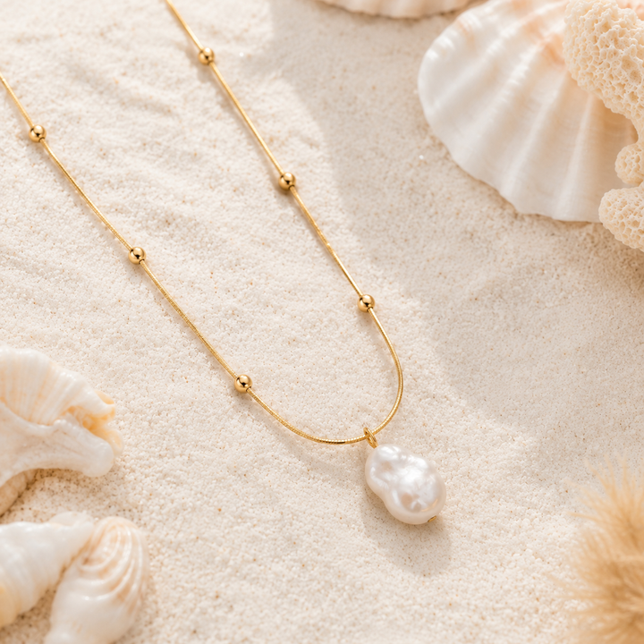 A delicate gold chain necklace with small gold beads and a baroque pearl pendant, styled on a sandy beach background with seashells.