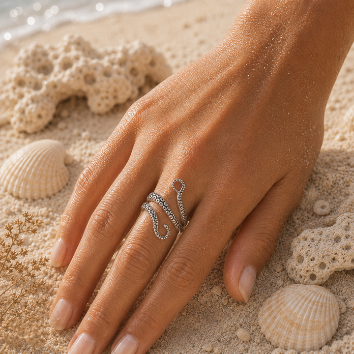 A close-up of a hand resting on sandy beach decor, wearing a silver octopus  ring. The background features seashells, coral, and sand.