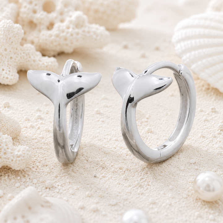 A pair of polished silver huggie hoop earrings shaped like whale tails, displayed on a sandy surface with white seashells and coral props.