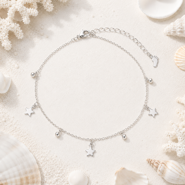 A delicate silver chain anklet with three small star charms and tiny bead accents, displayed on a light sandy surface surrounded by white seashells and coral.