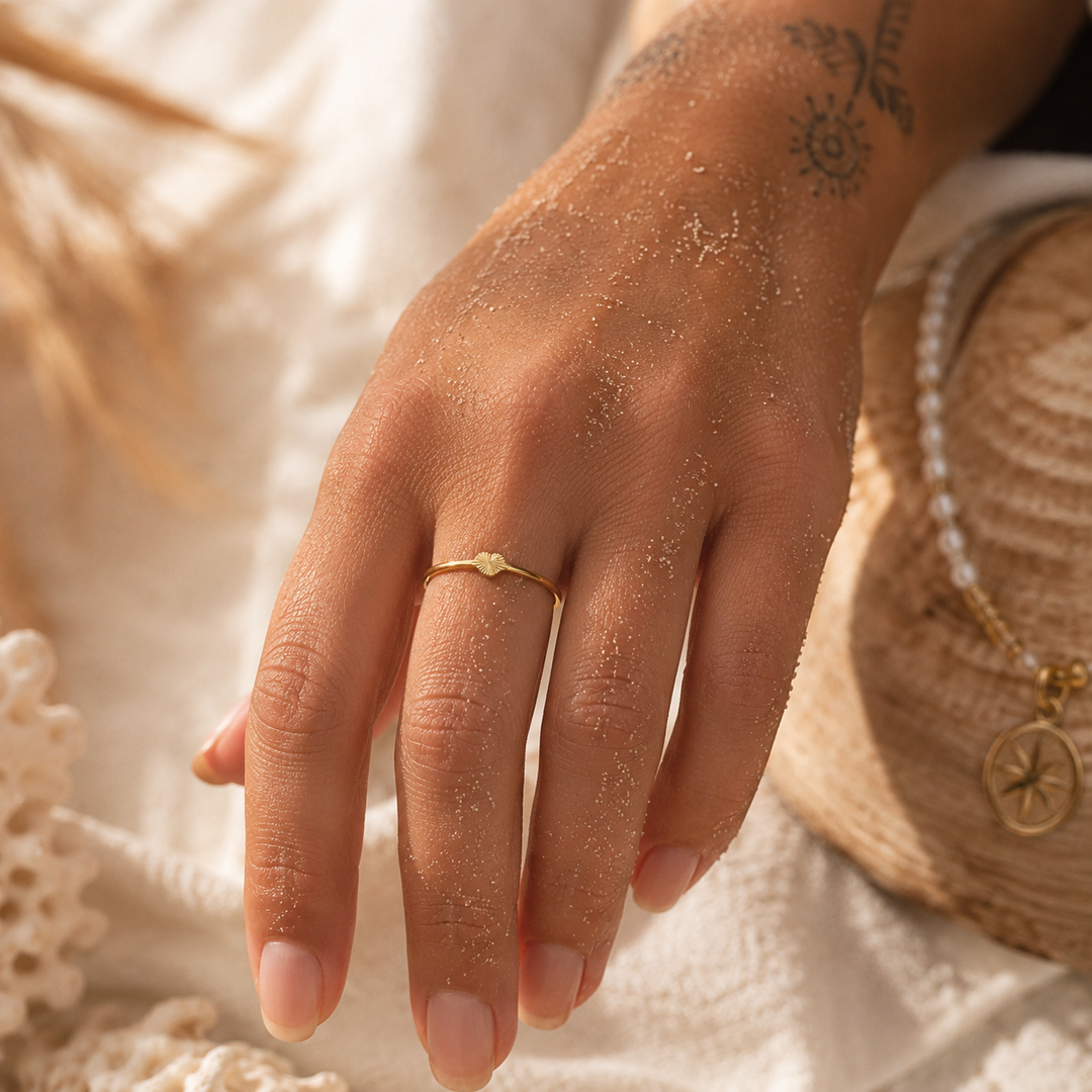 A close-up of a person's hand resting on a white textured surface, wearing a delicate gold ring with a small heart-shaped RING. 