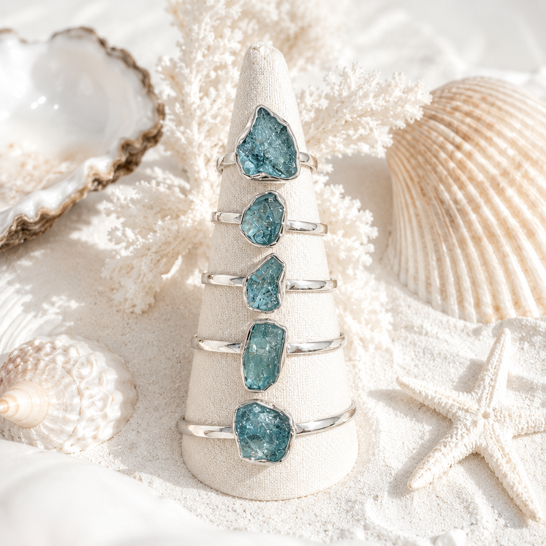 Raw aquamarine rings, displayed on a cream cone stand, styled on white sand with seashells and a starfish.