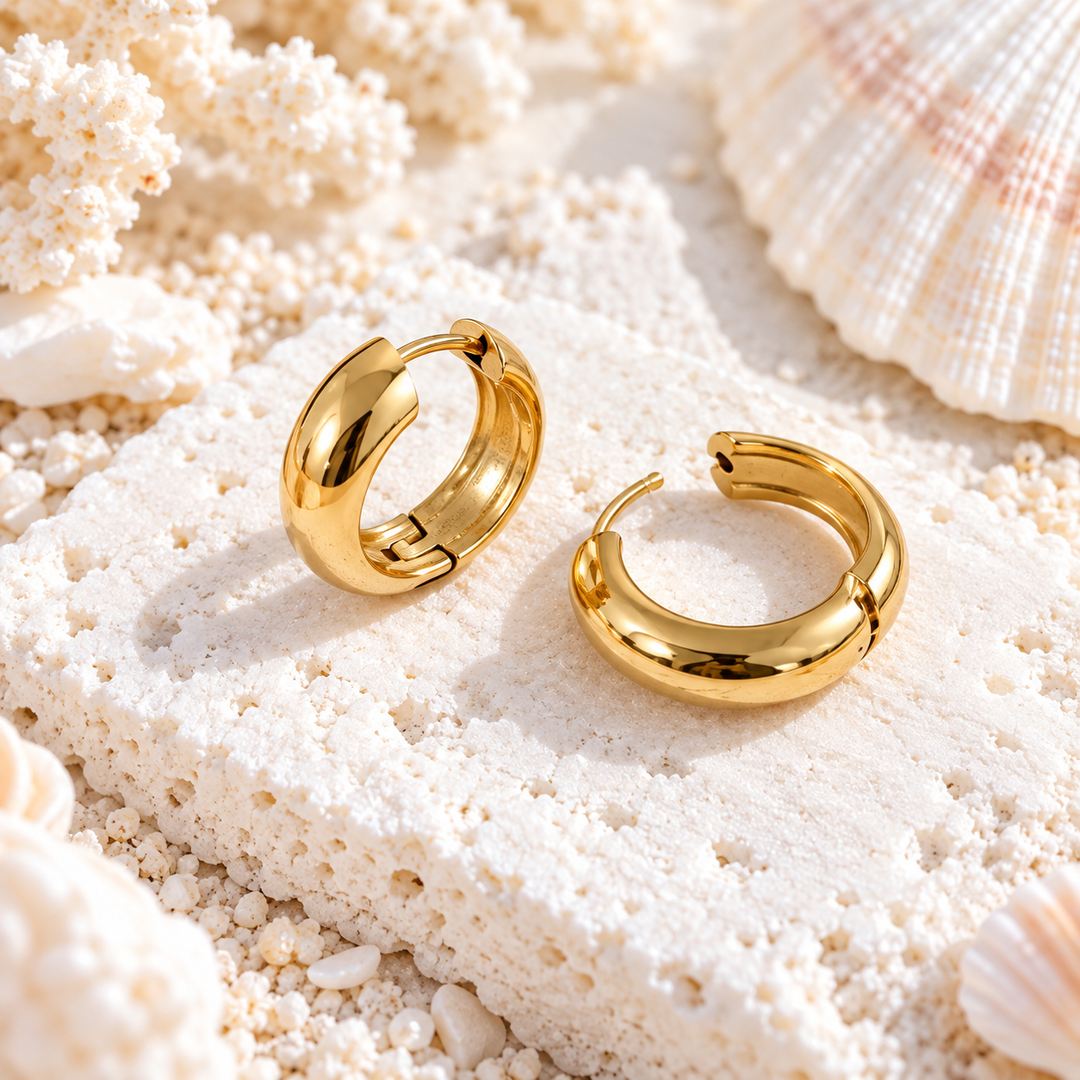 A pair of polished gold hoop earrings resting on a textured white stone slab surrounded by seashells and coral.