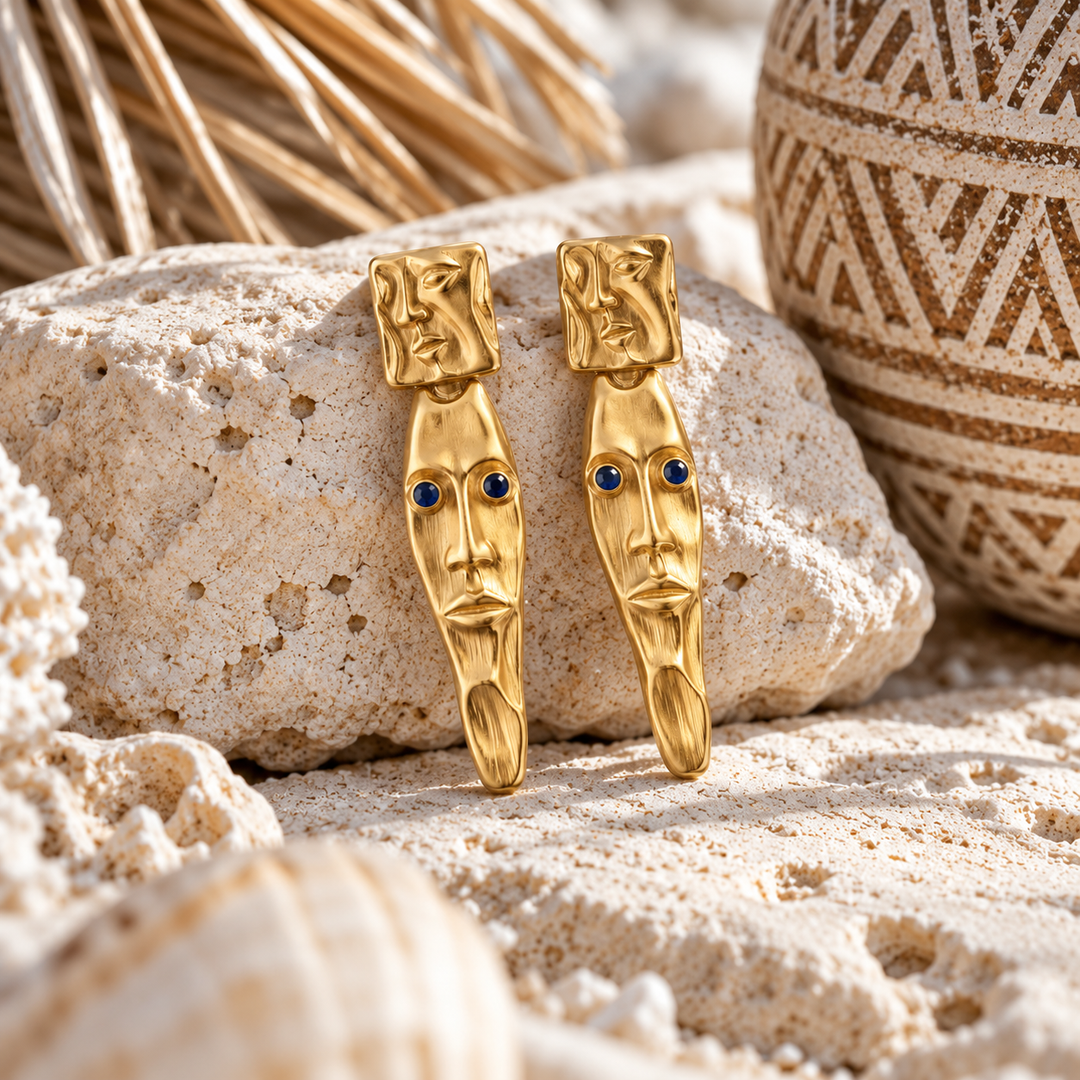 A pair of gold drop earrings shaped like stylized faces with small blue gemstone eyes, displayed on a textured beige rock with seashells and dried grass in the background.