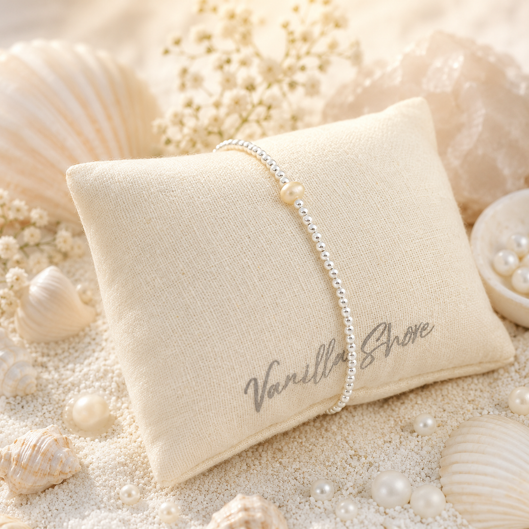 Pearl bracelet on a 'Vanilla Shore' pillow surrounded by seashells and pearls.