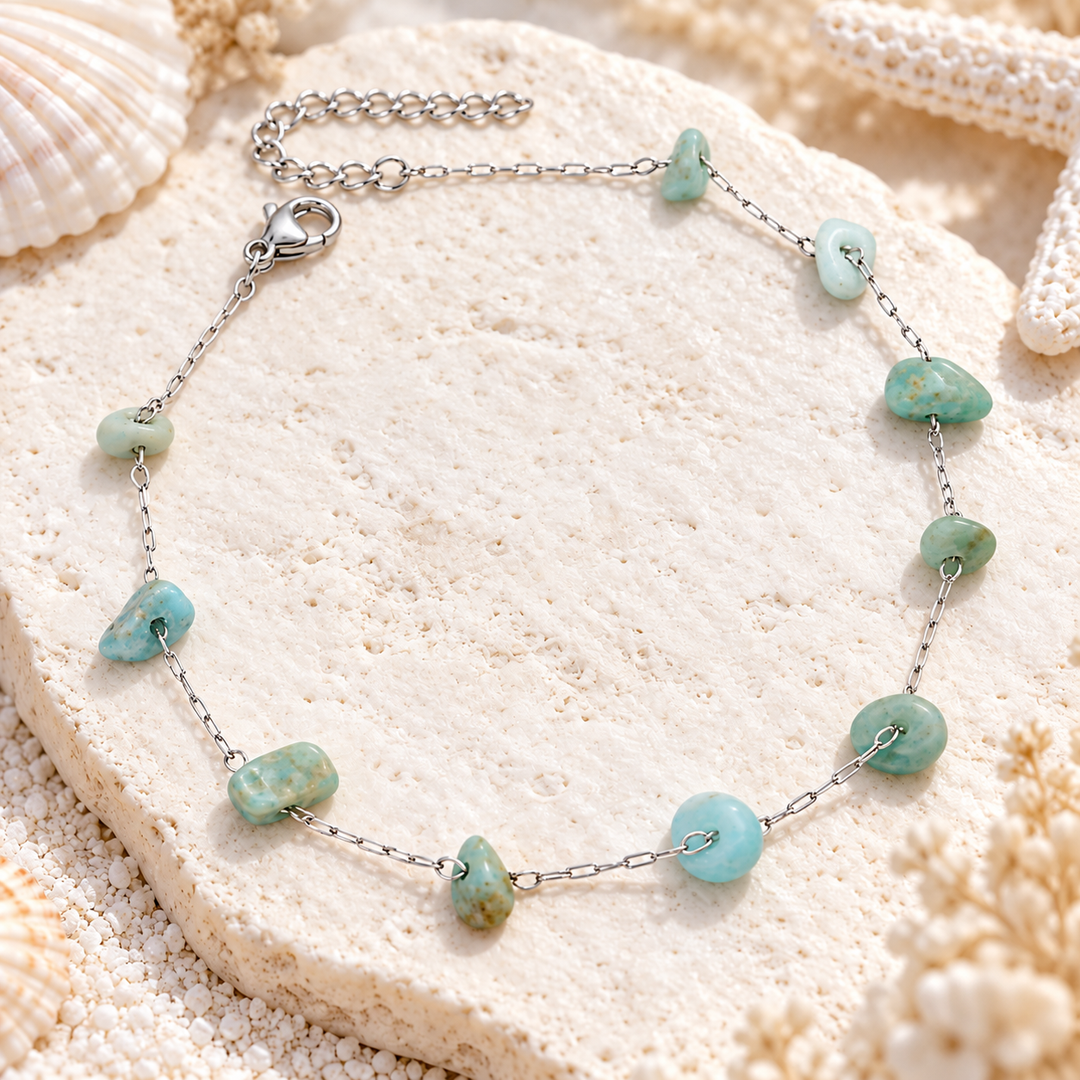 A silver chain anklet with irregular light blue-green stone beads, arranged in a circle on a textured beige stone with seashell and coral props.